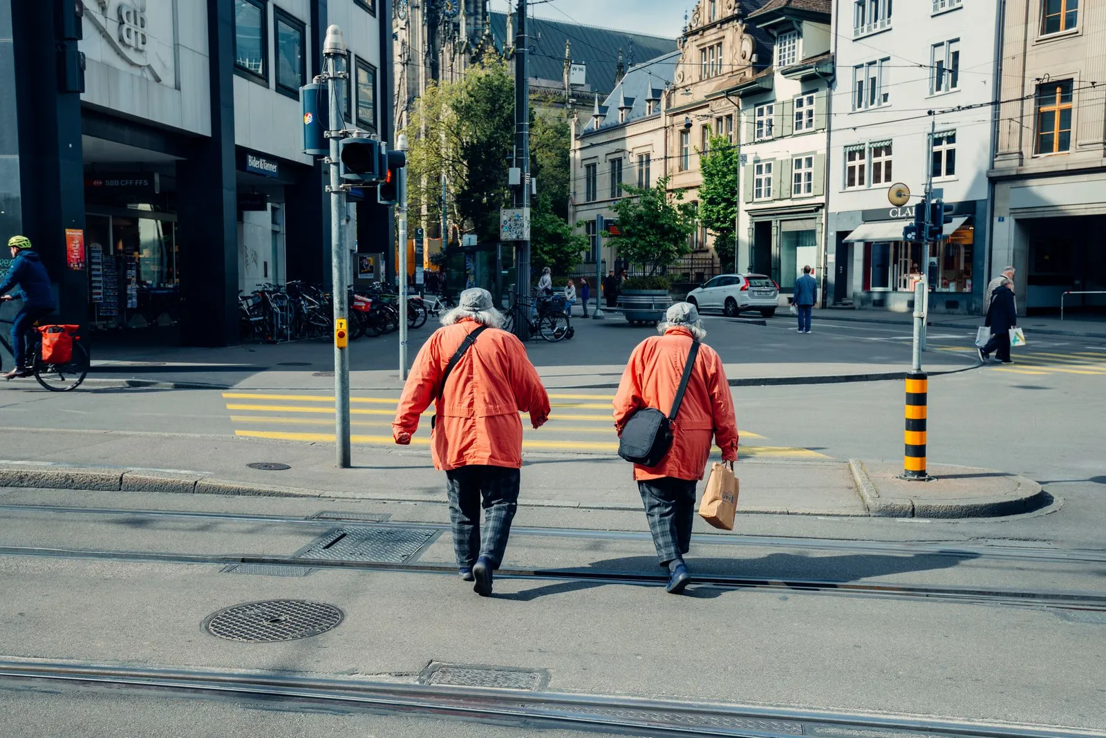 Elderly couple in matching red jackets crossing a street in the vibrant city of Basel, Switzerland.