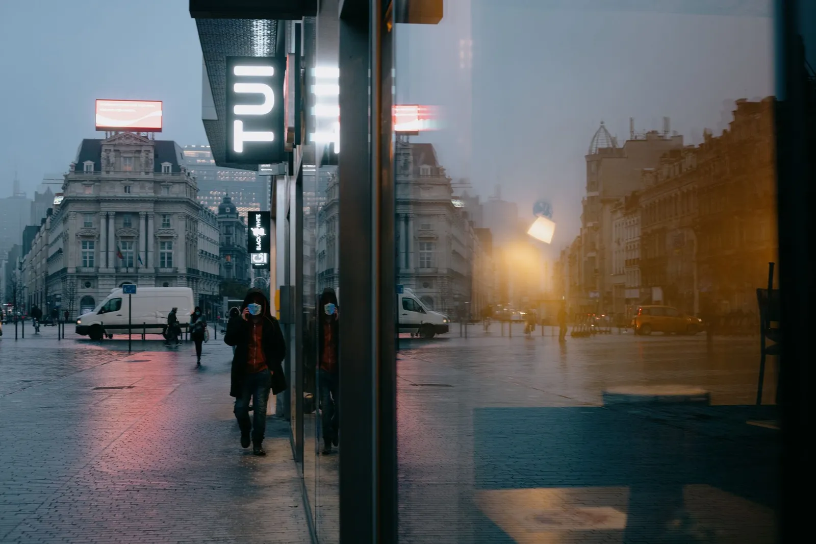 Anonymous people in medical masks walking on Place de Brouckere square near modern building with glass walls in rainy evening during coronavirus in Brussels