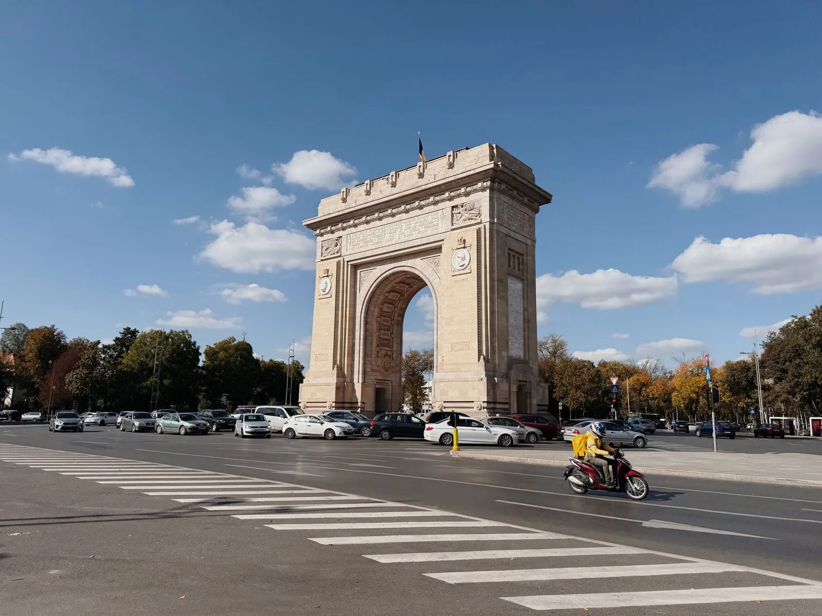 Triumphal arch in Bucharest against a blue sky, with traffic passing by.