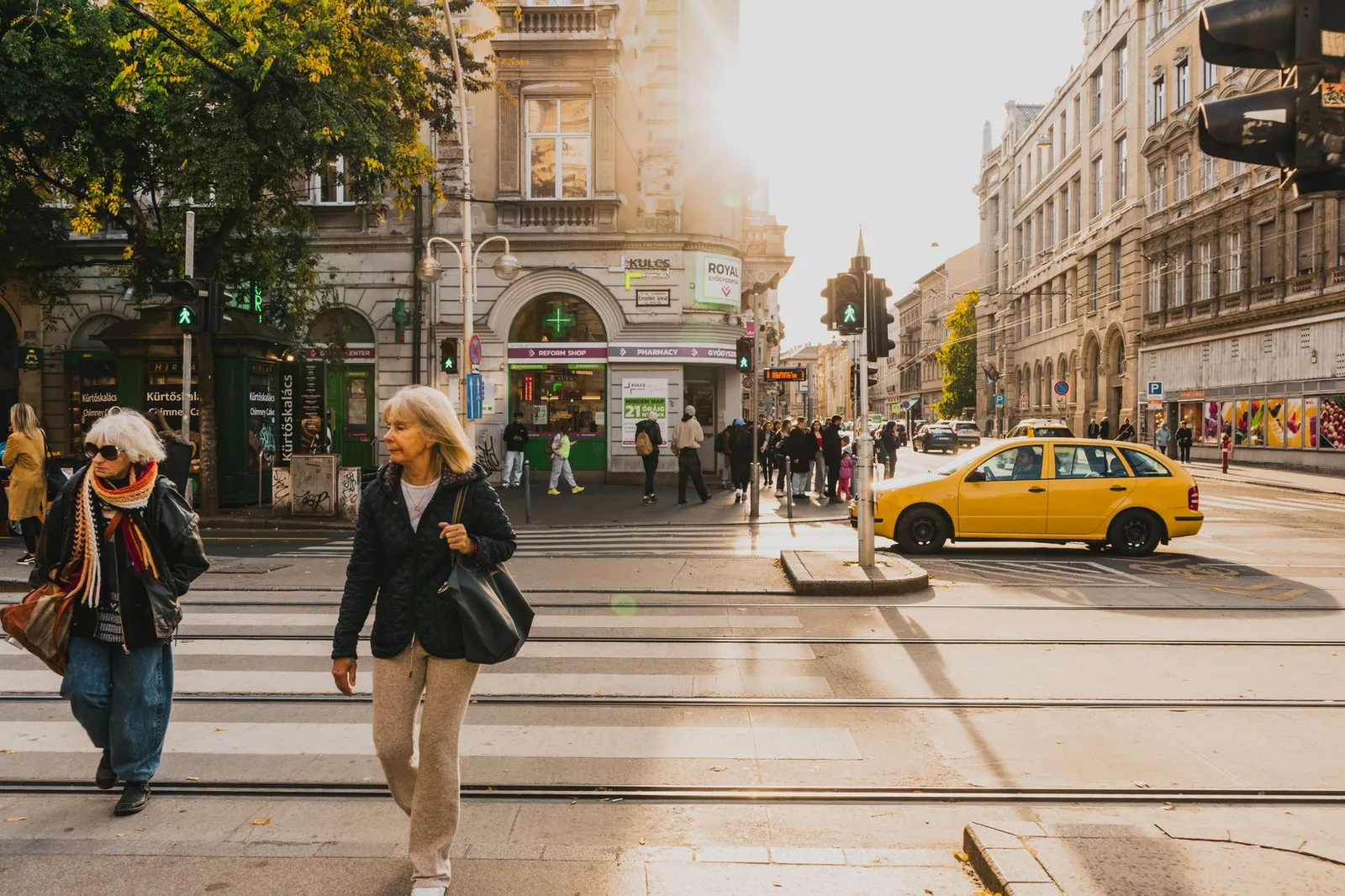 A bustling street intersection in Budapest with pedestrians and a yellow taxi under soft sunlight.