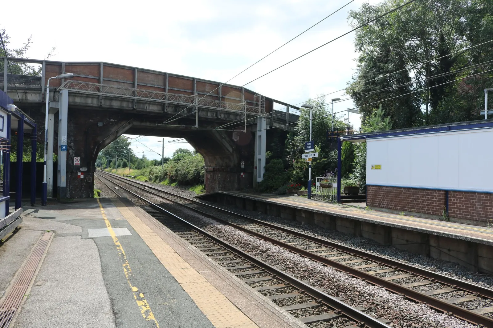 Empty railway station in Holmes Chapel, England with tracks and bridge on a sunny day.