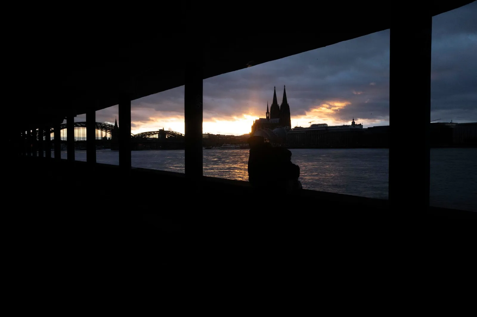 A serene sunset view of Cologne Cathedral and the Rhine River framed by a colonnade silhouette.