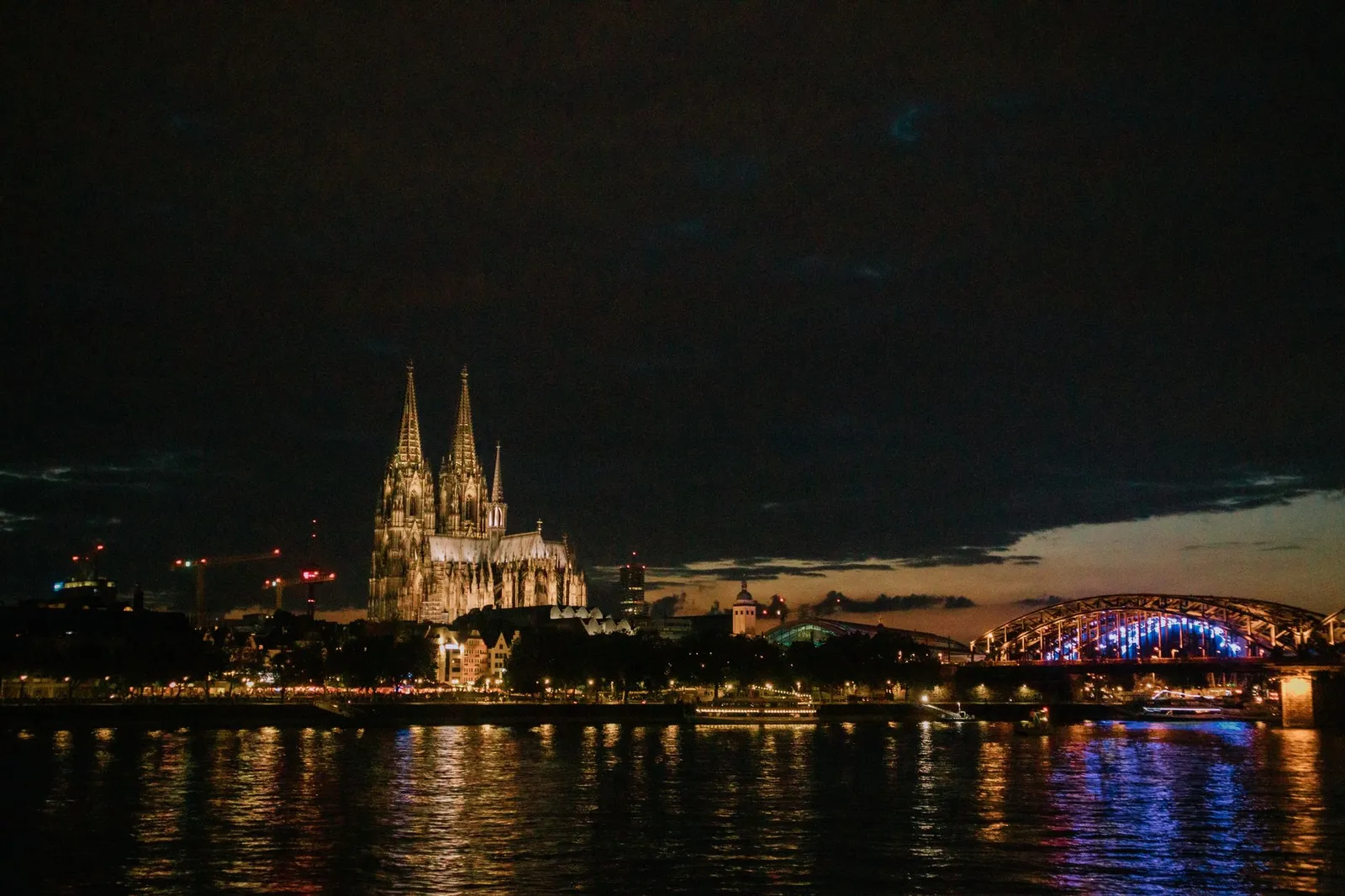 View of Cologne Cathedral and Hohenzollern Bridge illuminated at night, reflecting in the Rhine River