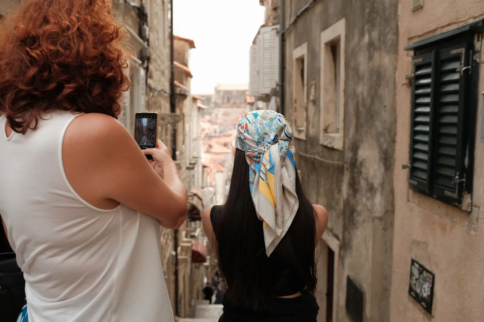 Two women explore a narrow alley in Dubrovnik's historic old town, capturing the moment on a smartphone.