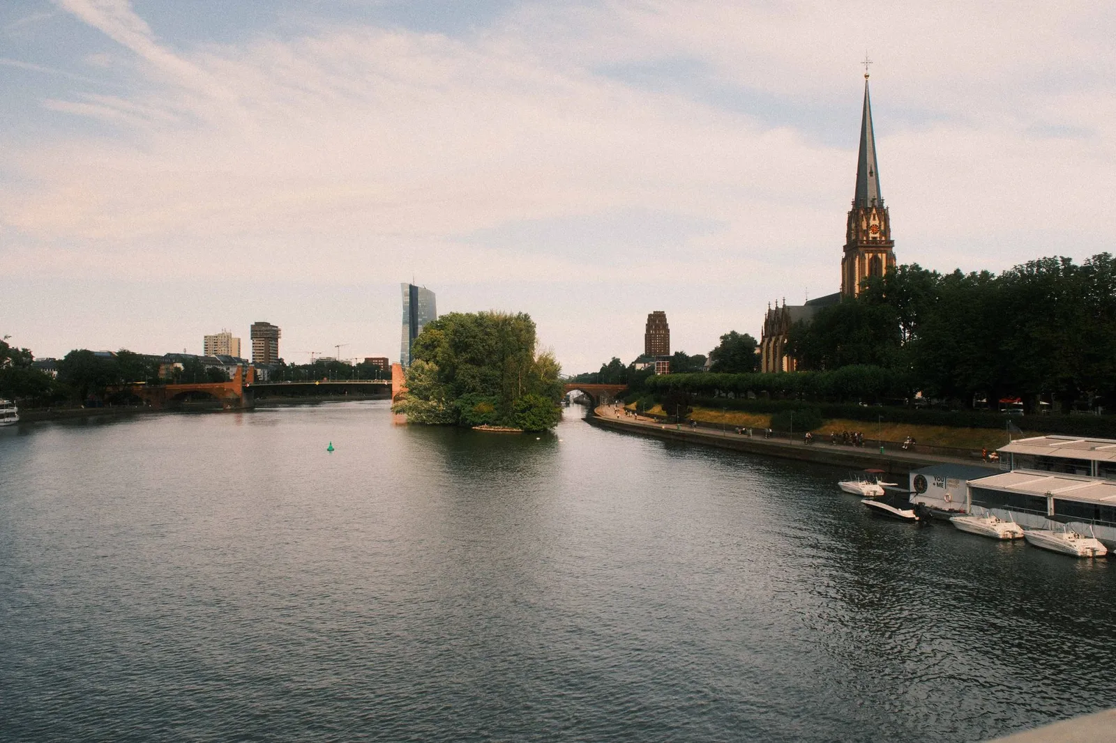 Peaceful river scene in Frankfurt with cathedral, boats, and skyline under a moody sky.