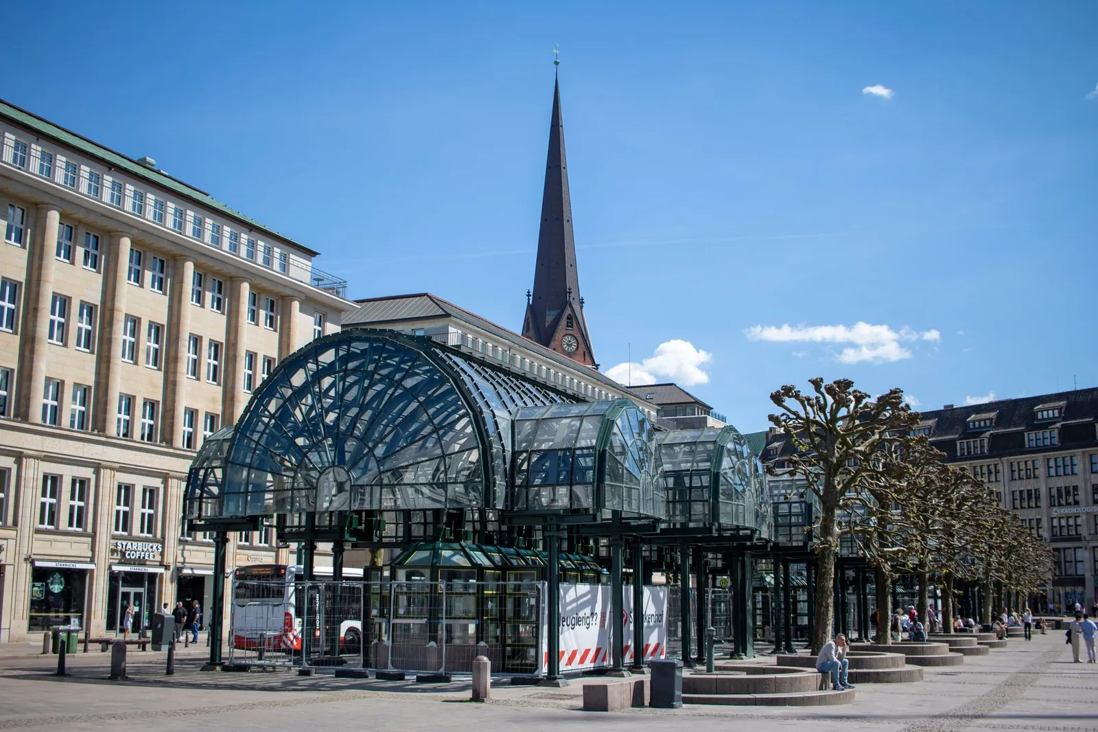 Glass-covered bus stop at Rathausmarkt, Hamburg, with historic buildings under a clear blue sky.