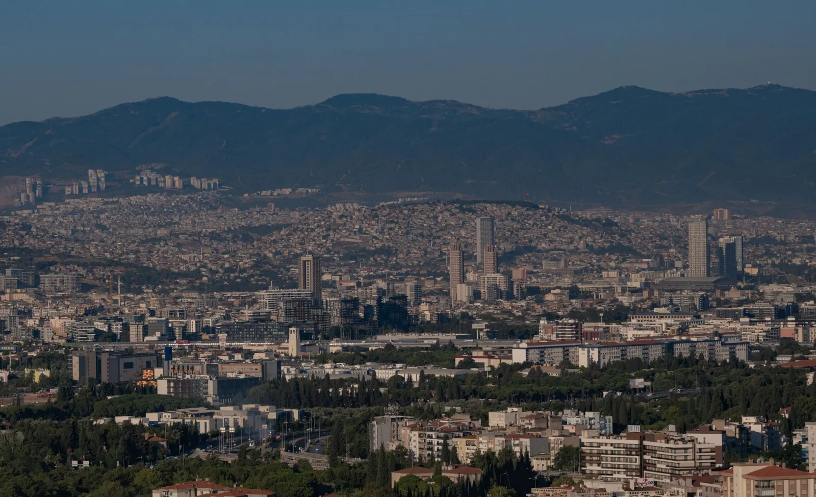 Aerial view of Izmir, Türkiye, showcasing cityscape and mountainous backdrop under a clear blue sky.