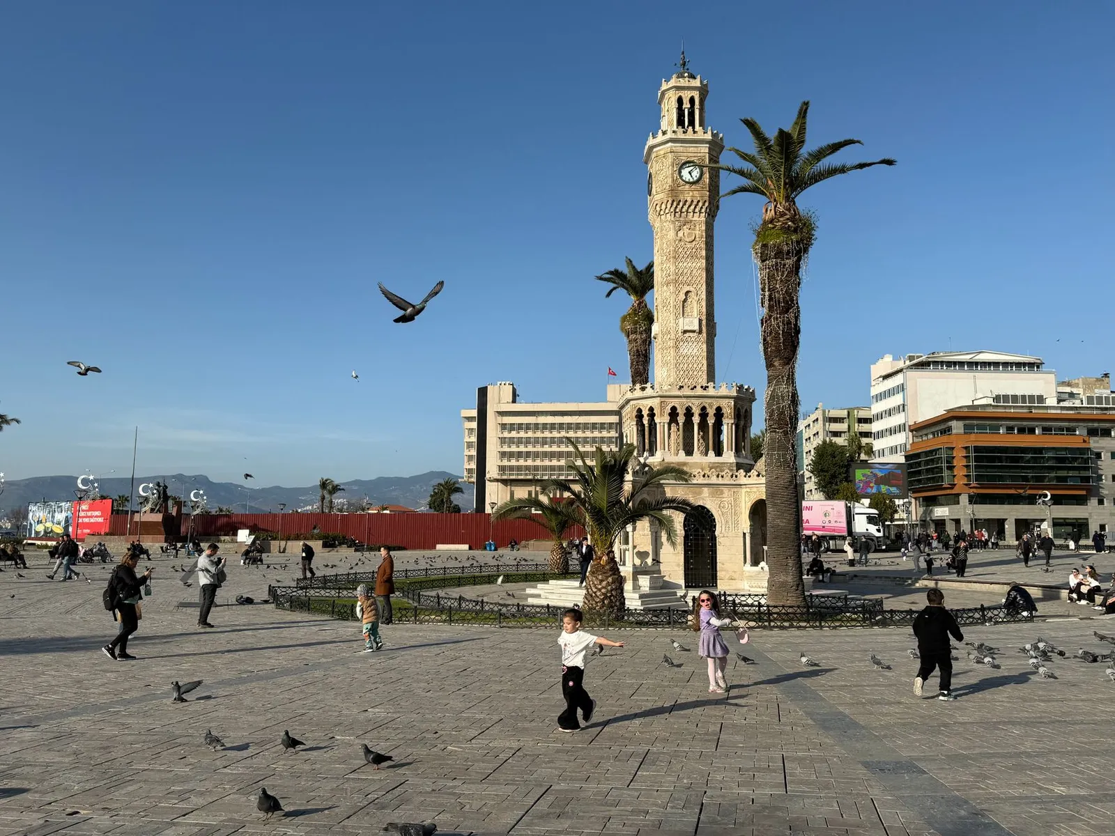Scenic view of İzmir's iconic Clock Tower in Konak Square with playful children and vibrant pigeons.