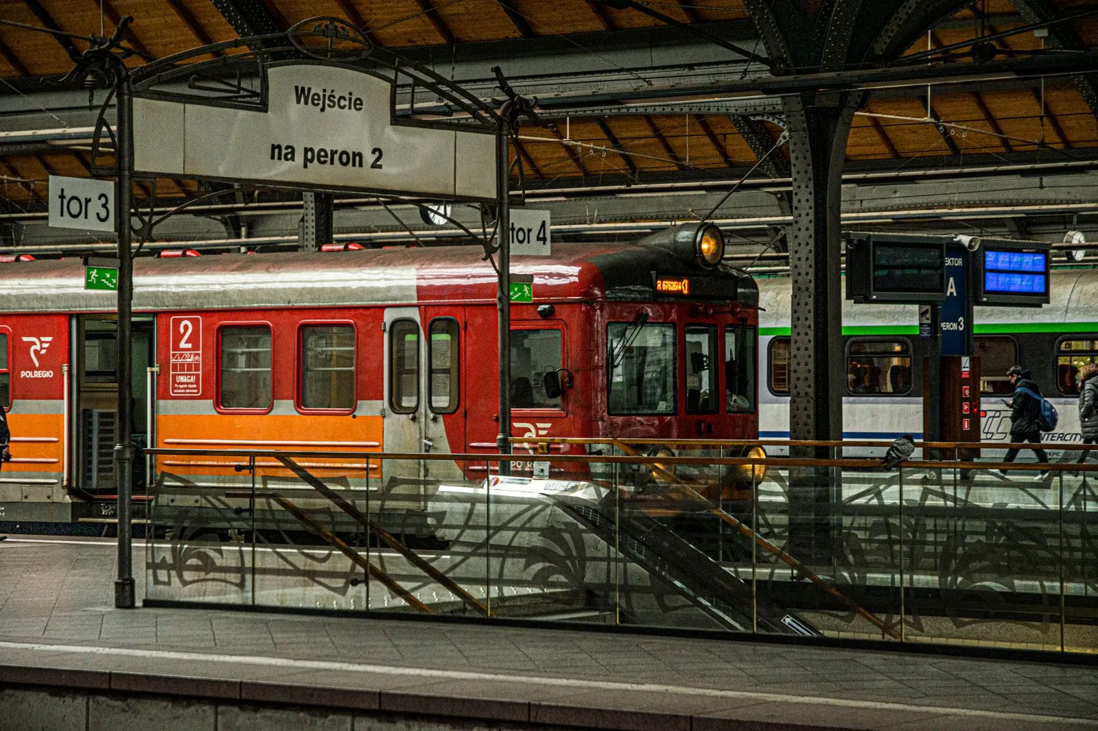 A vibrant red train awaits departure at a historic Polish train station, platform signs visible.