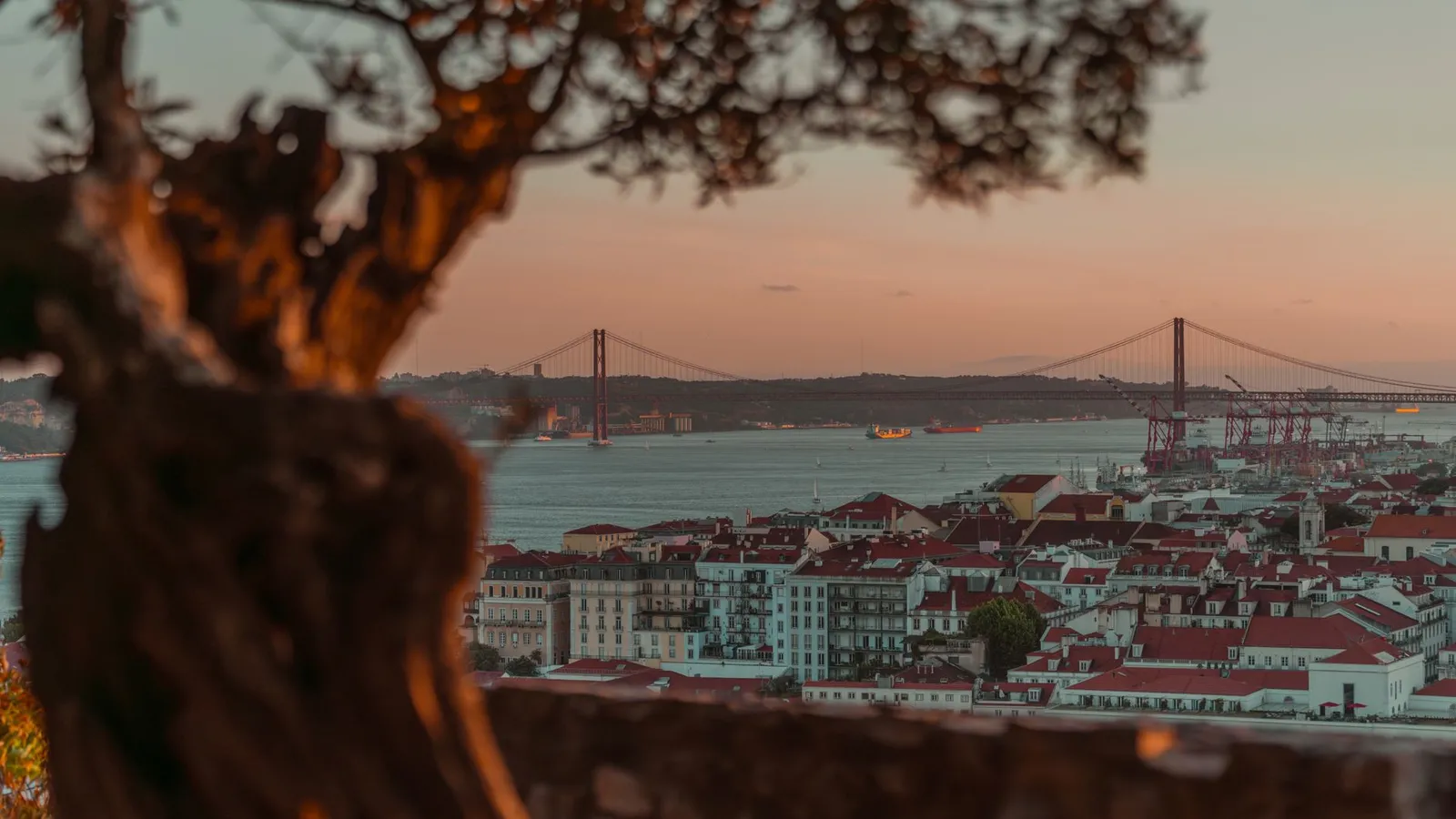 Beautiful sunset over Lisbon with a view of the iconic 25 de Abril Bridge, framed by trees.