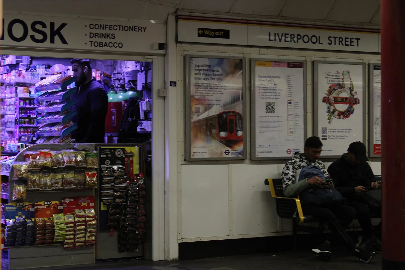 Night scene at Liverpool Street Station with a vibrant kiosk and waiting passengers.