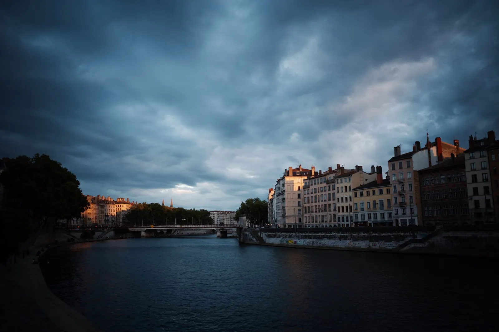 Historic buildings line the river under dramatic clouds, creating a moody urban landscape.