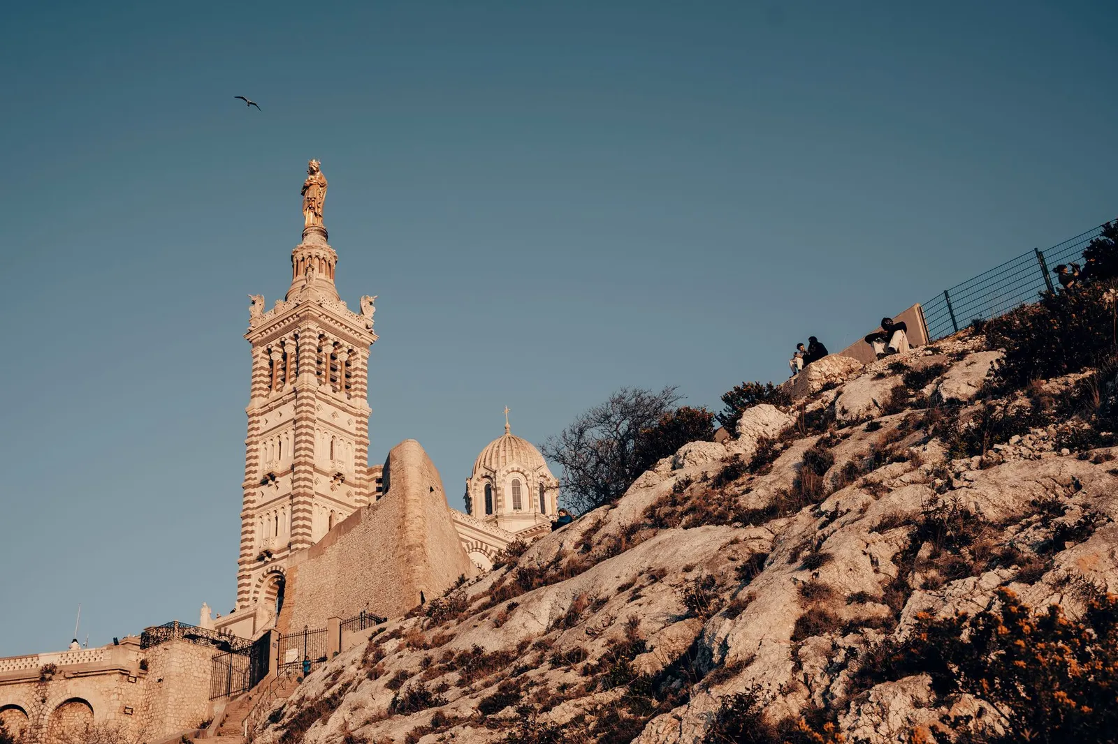 View of Notre-Dame de la Garde with rocky cliff in Marseille under bright summer sky.