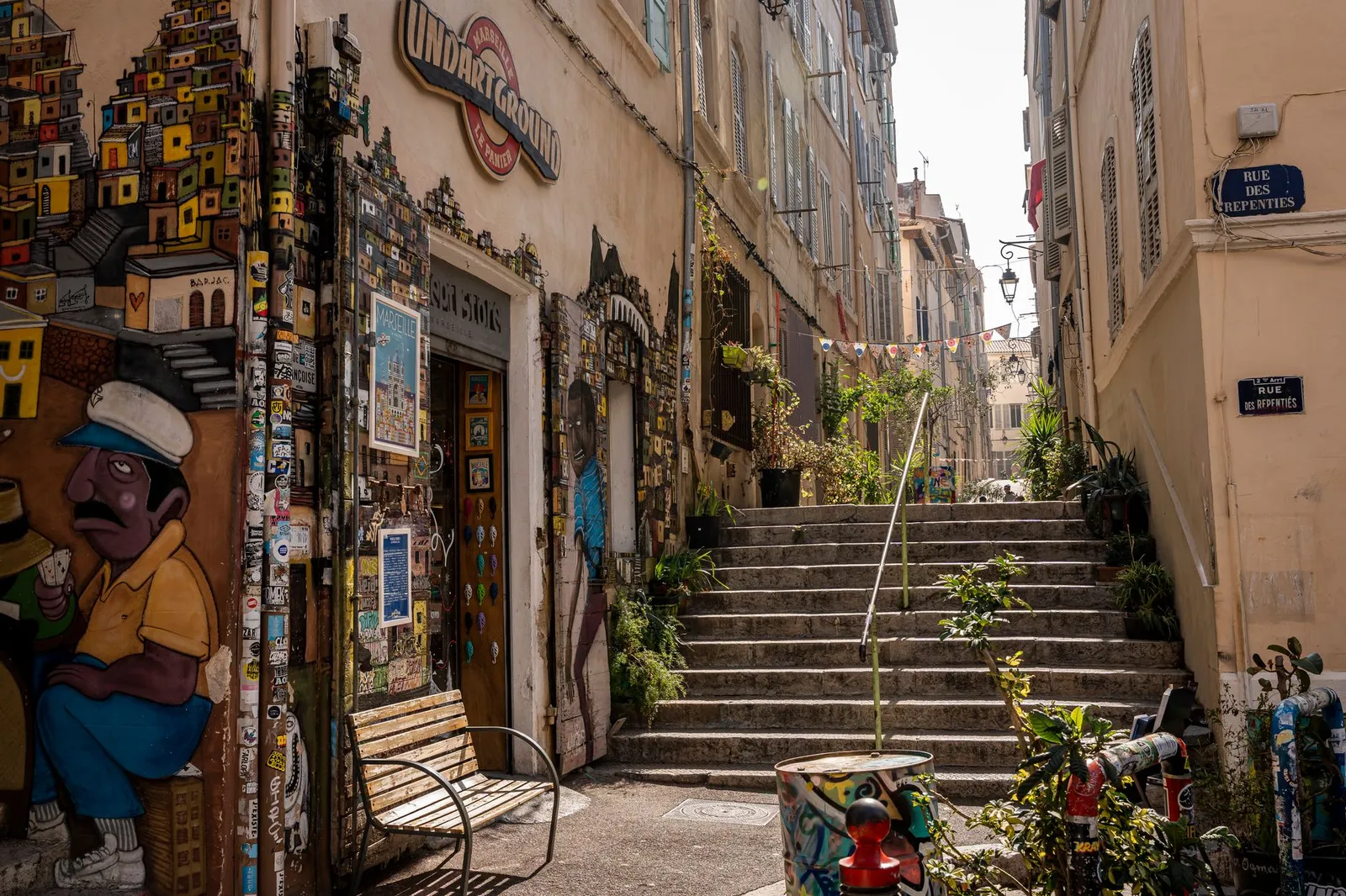 Sunlit narrow street in Marseille adorned with vibrant murals and lush plants on a bright day.
