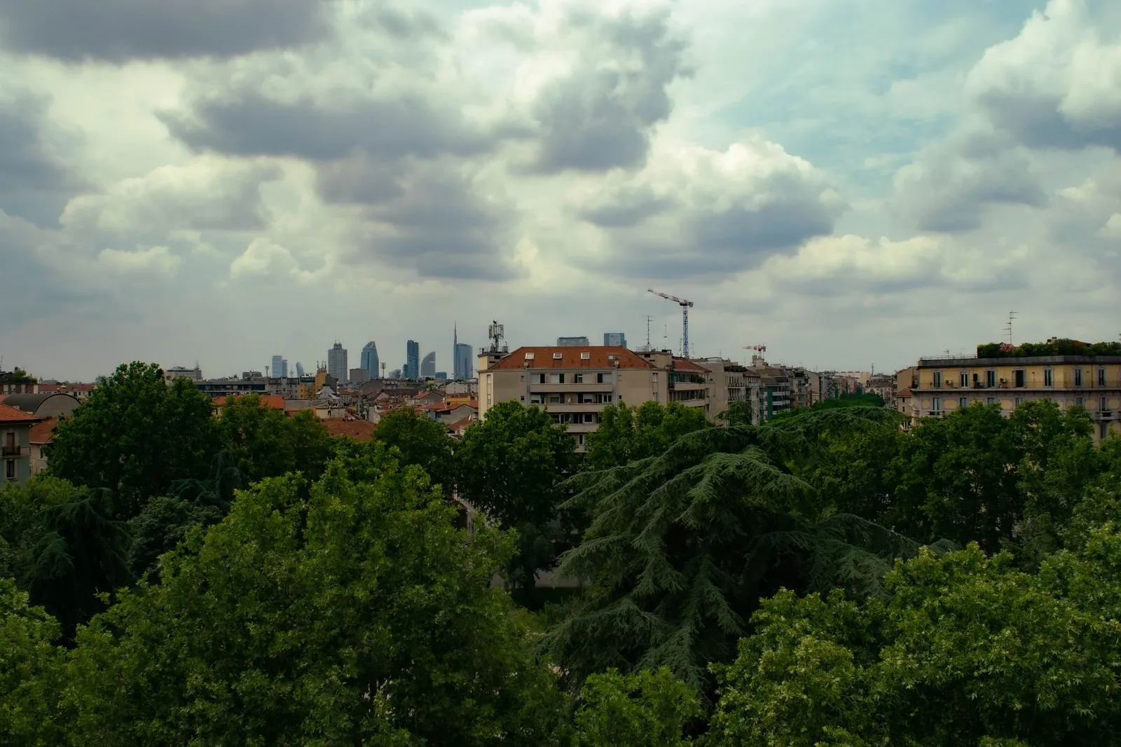 Skyline of Milan with clear view of urban buildings amidst lush green trees under a cloudy sky.