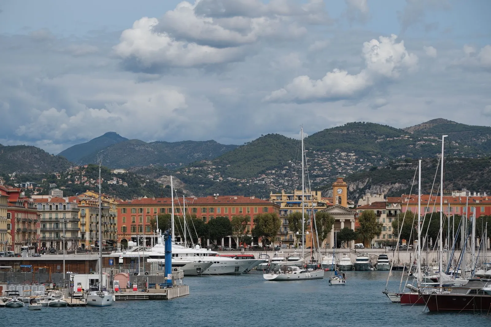 Scenic view of Port Lympia in Nice, France, featuring yachts, mountains, and classic architecture.