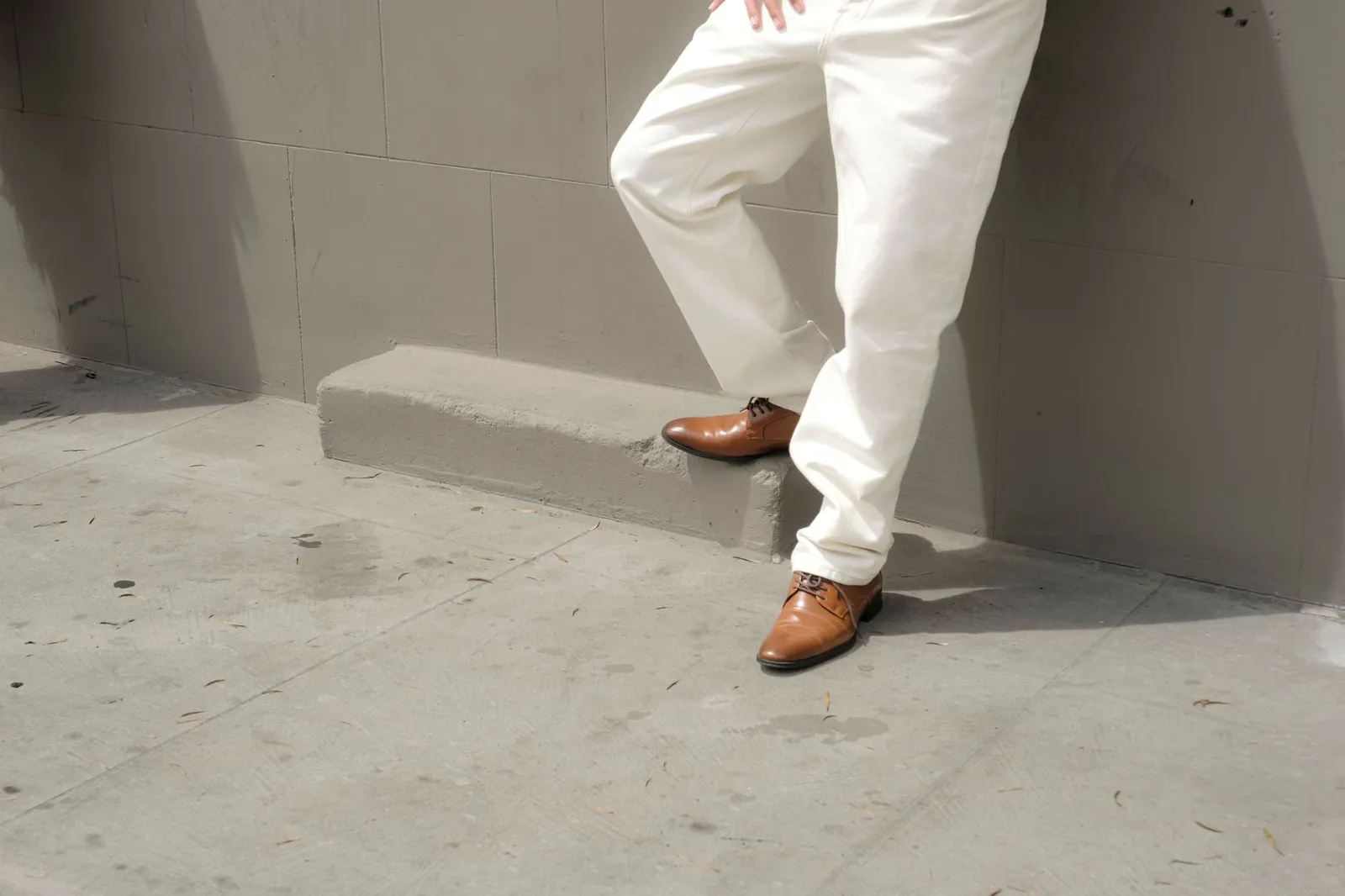 A person wearing classic brown Oxfords and white pants stands stylishly against a concrete urban wall.