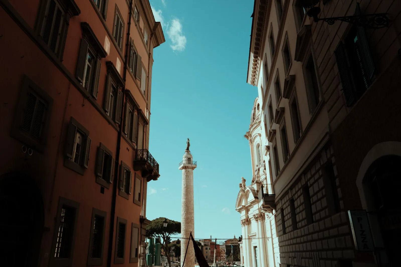 Dramatic view of Trajan's Column framed by historic buildings in Rome, Italy under a clear blue sky.