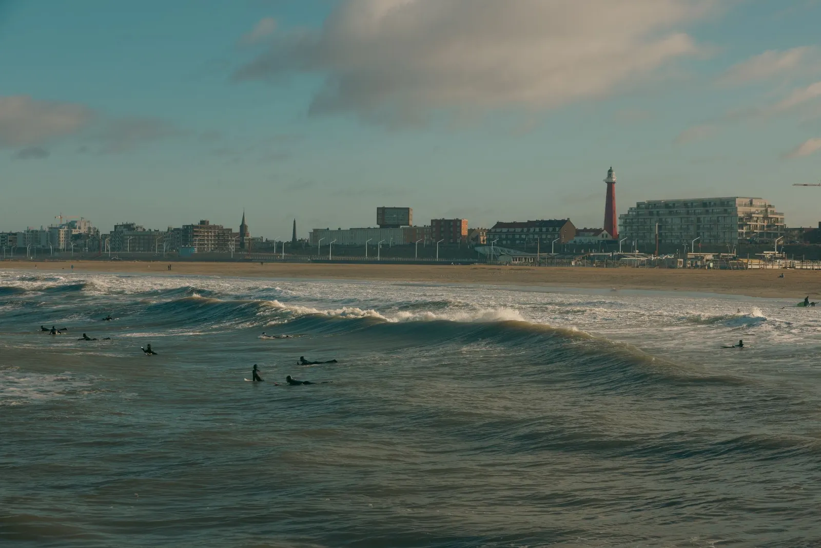 Surfers ride waves near the beach in The Hague, Netherlands with the city skyline as a backdrop.