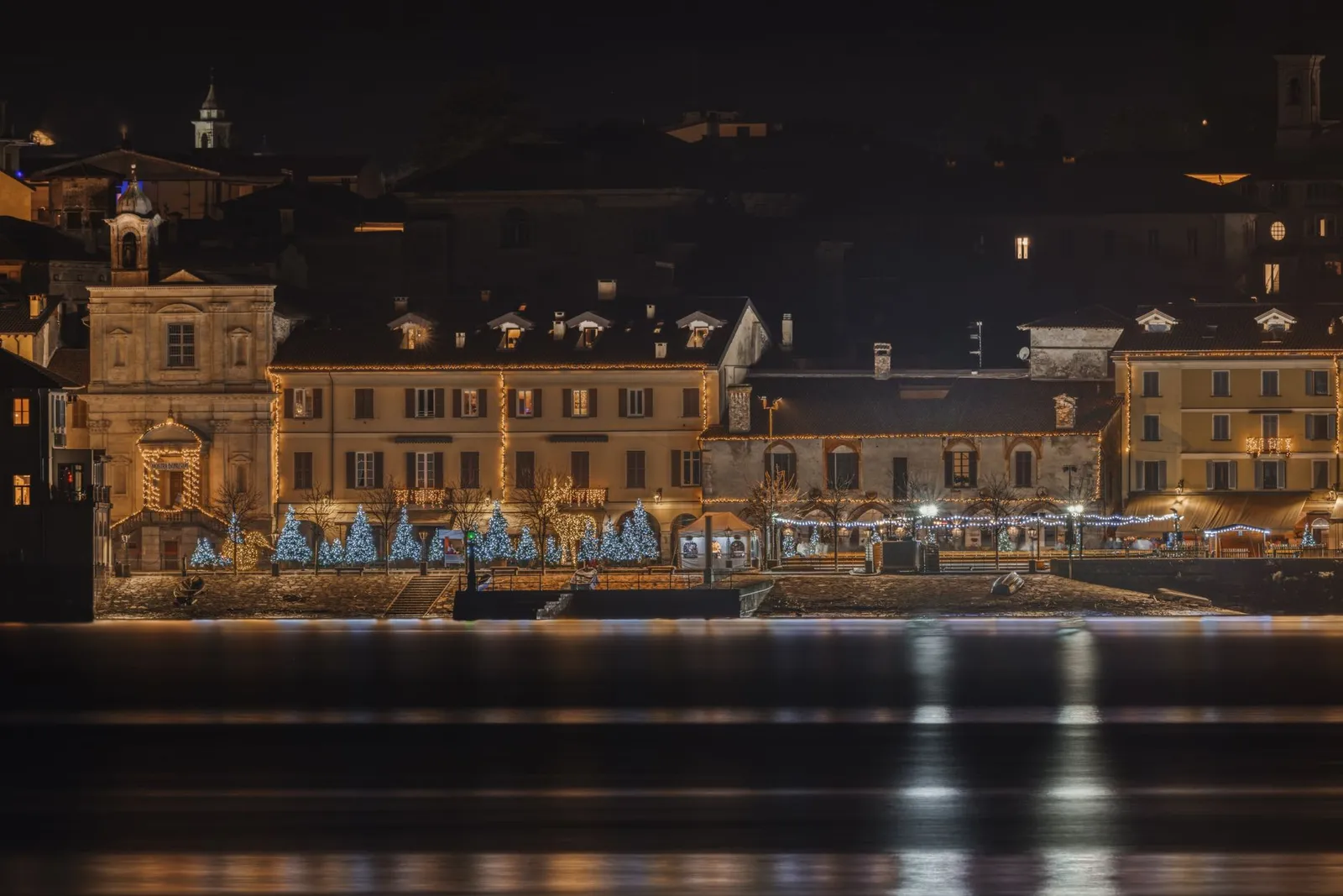 Beautiful riverside scene with holiday lights on buildings at night, reflecting on water.