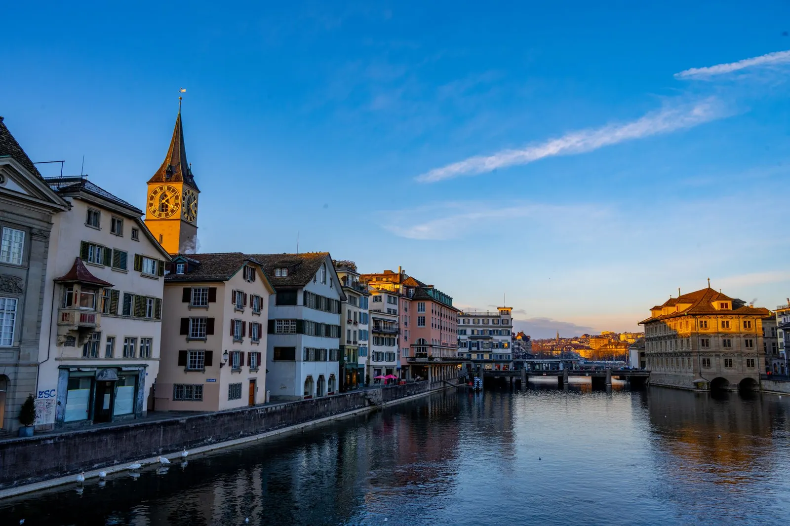 Beautiful view of Zurich cityscape along the Limmat River featuring St. Peter's Church at sunset.