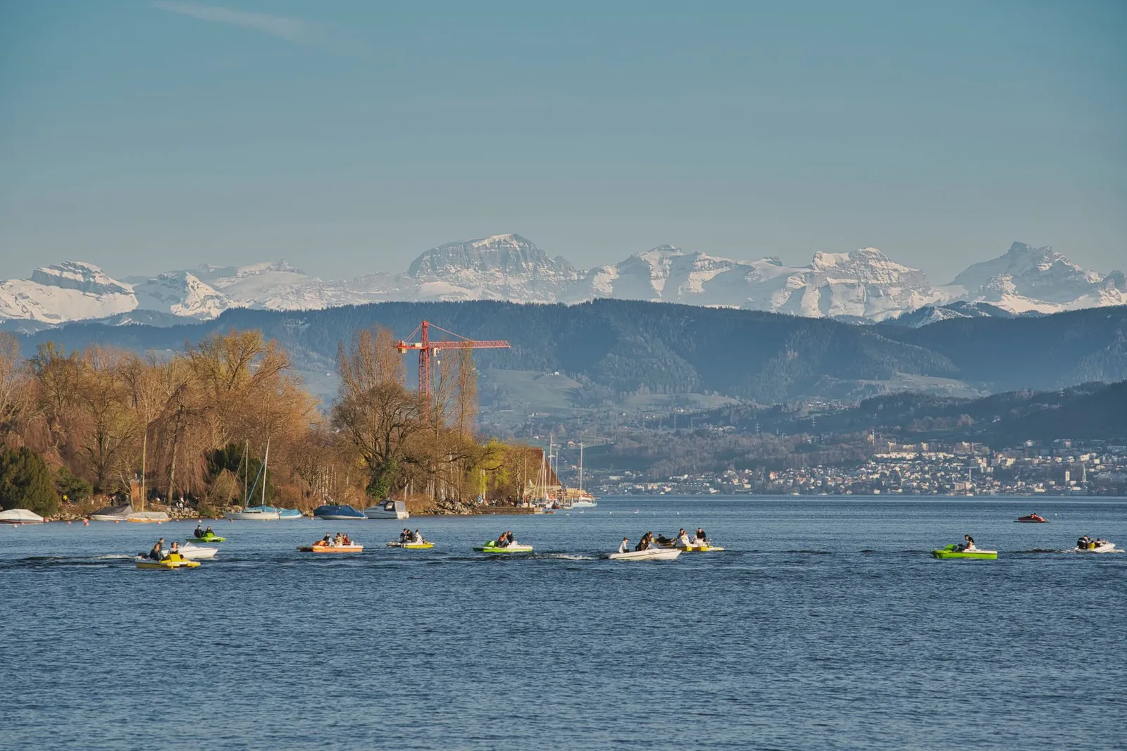 A picturesque view of Lake Zurich with people boating and the snowy Alps in the background.